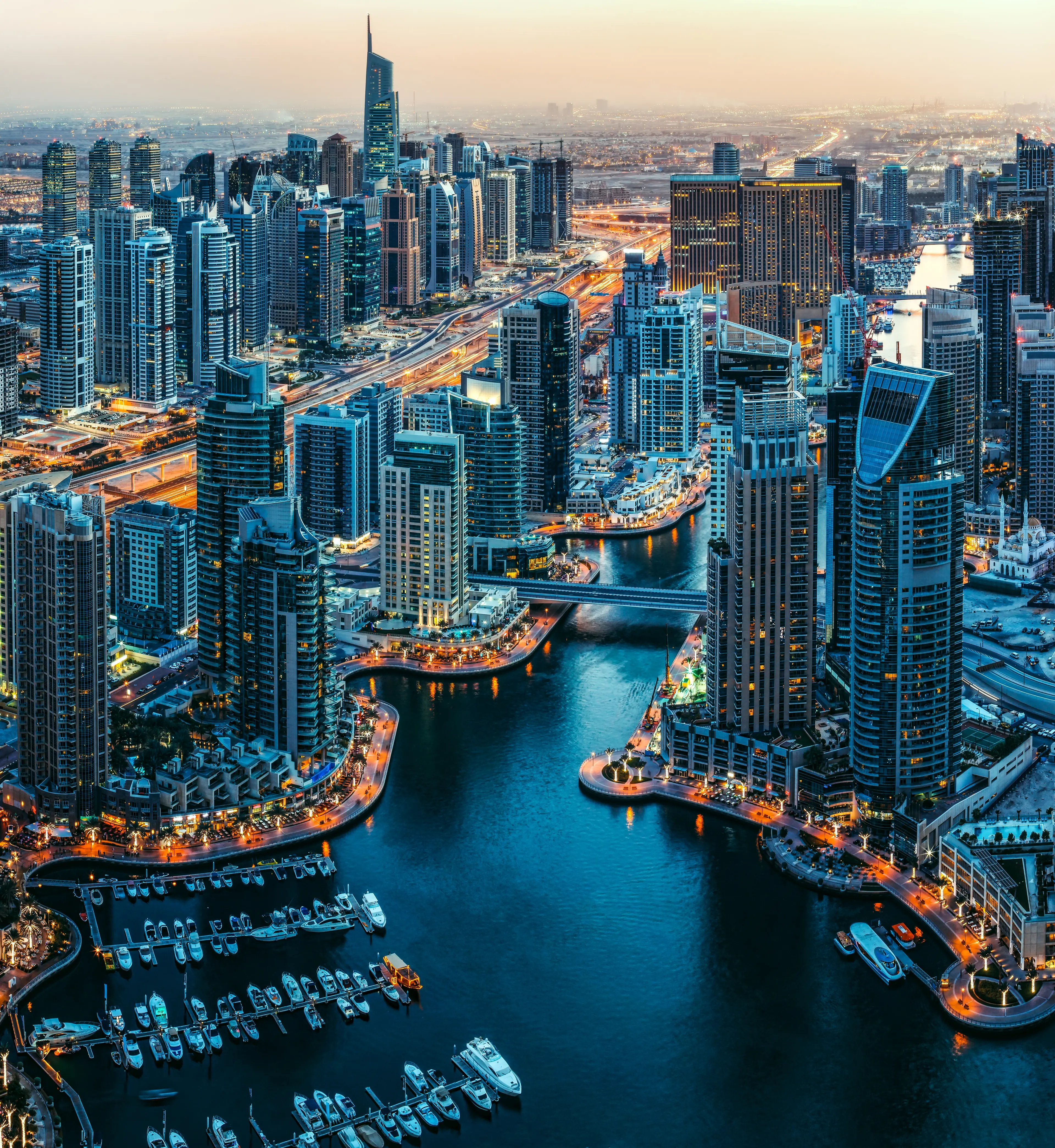 Aerial view of Dubai Marina at dusk with illuminated skyscrapers and yachts in the waterway.